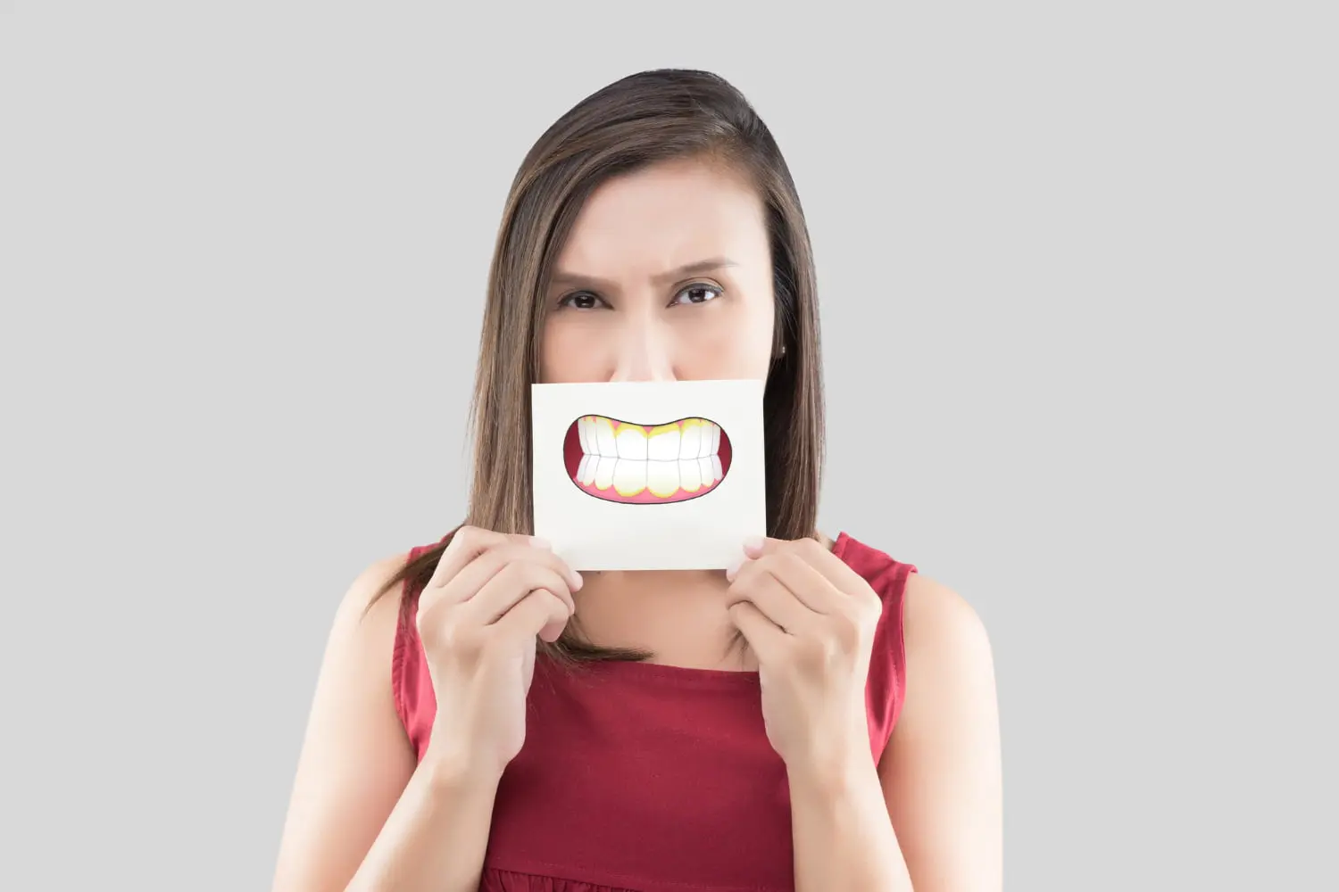 A woman playfully holds a paper with a cartoon smile featuring an underbite over her mouth against a gray background at Levine Orthodontics in Fort Myers, Cape Coral or Lehigh Acres, FL.