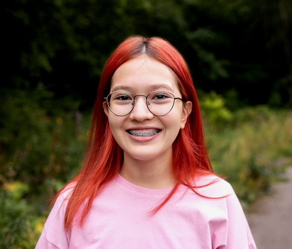 Smiling outdoors on a path surrounded by greenery, a person with red hair, glasses and metal braces showcases the results of Levine Orthodontics in Fort Myers, Cape Coral or Lehigh Acres, FL.