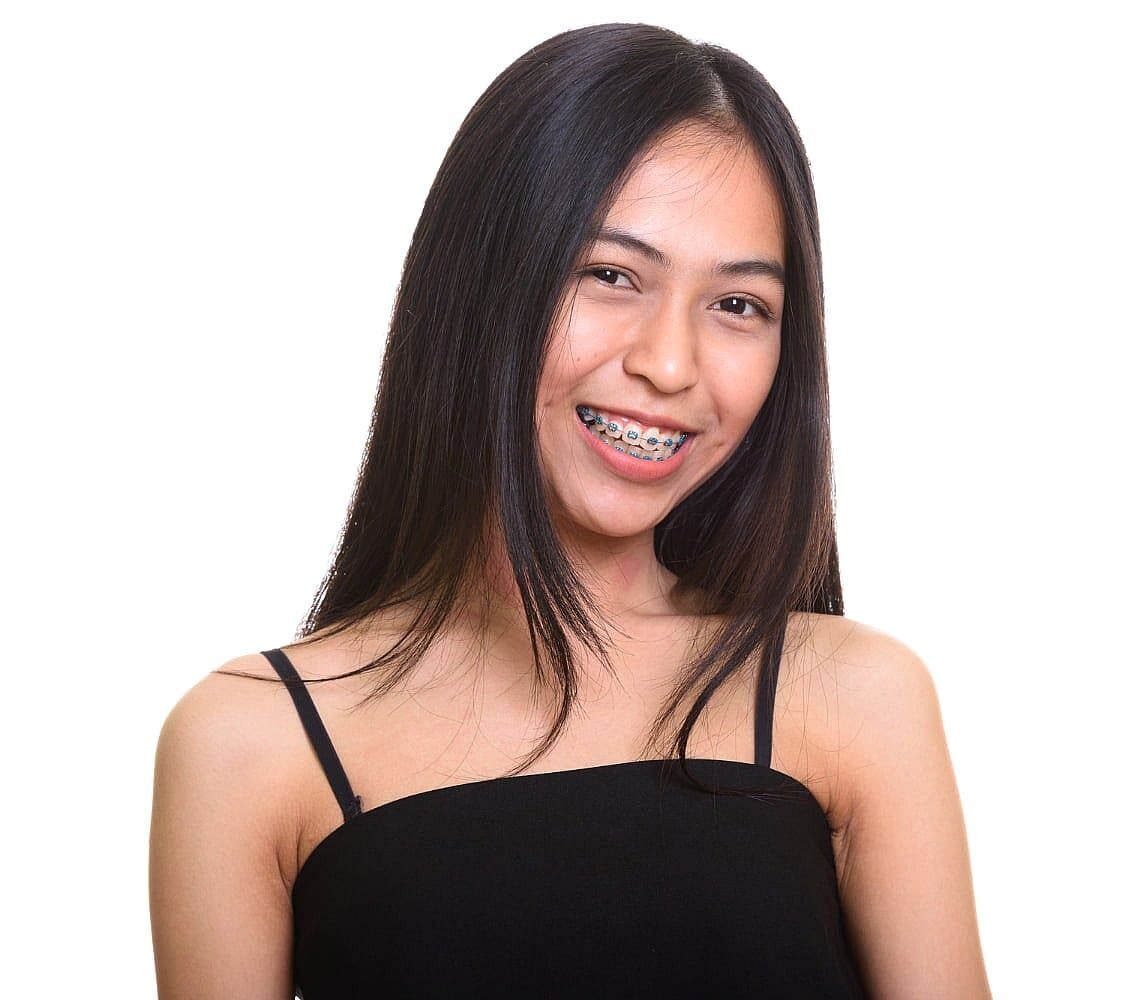 A young woman with long dark hair and metal braces, wearing a sleeveless black top, smiles at the camera against a white background—an example of the confident results achieved with Levine Orthodontics in Fort Myers, Cape Coral or Lehigh Acres, FL.