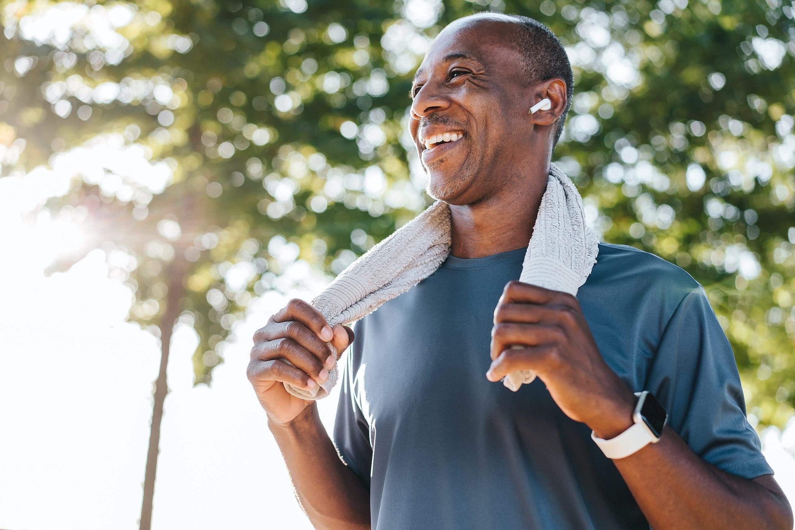 Smiling outdoors with trees and sunlight in the background, a mouth breather enjoys a refreshing workout session—man in athletic wear, towel around his neck, and earbuds in—at Levine Orthodontics in Fort Myers, Cape Coral or Lehigh Acres, FL.